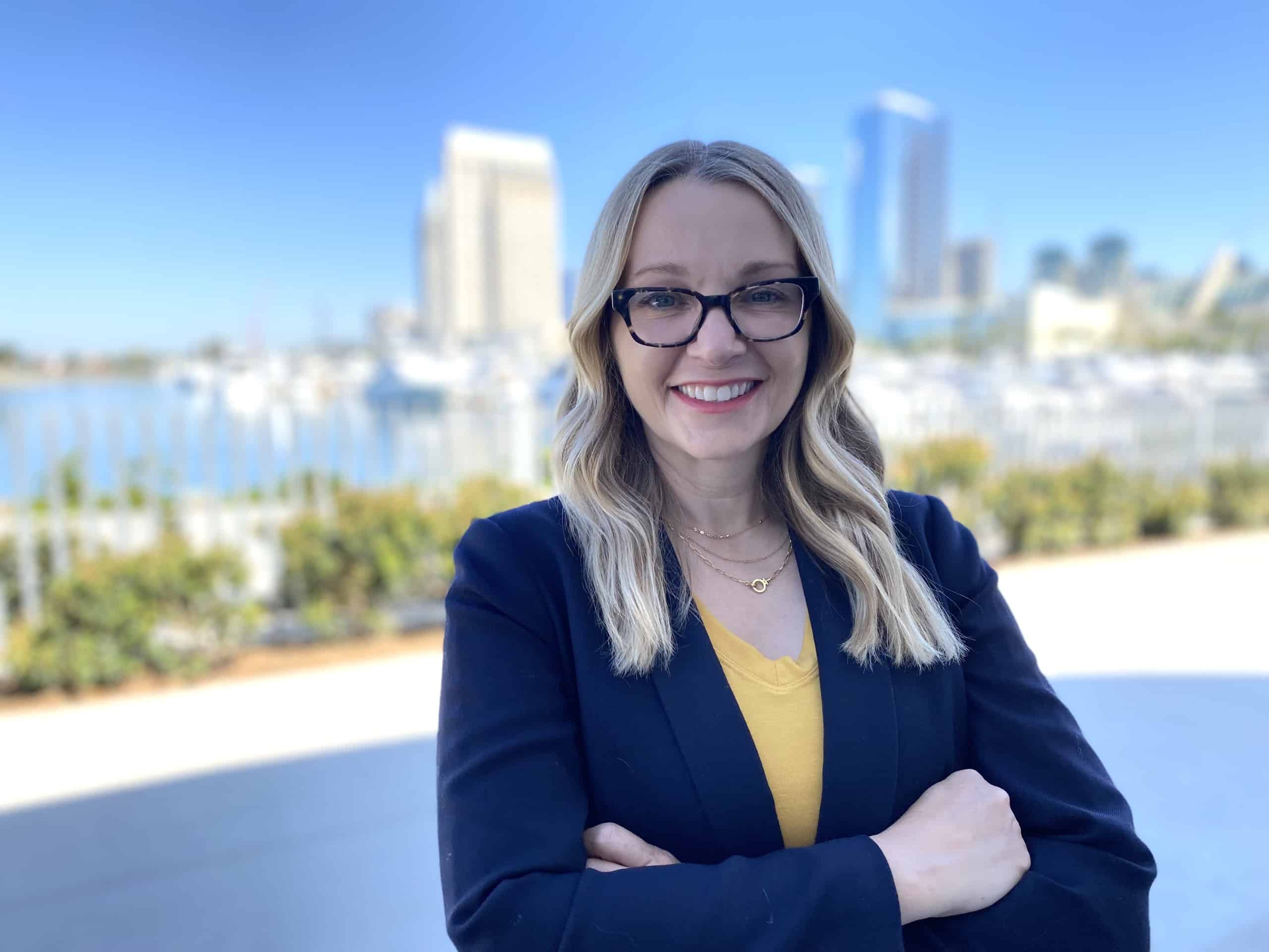 A blond woman in glasses stands smiling with her arms crossed in front of the port of San Diego