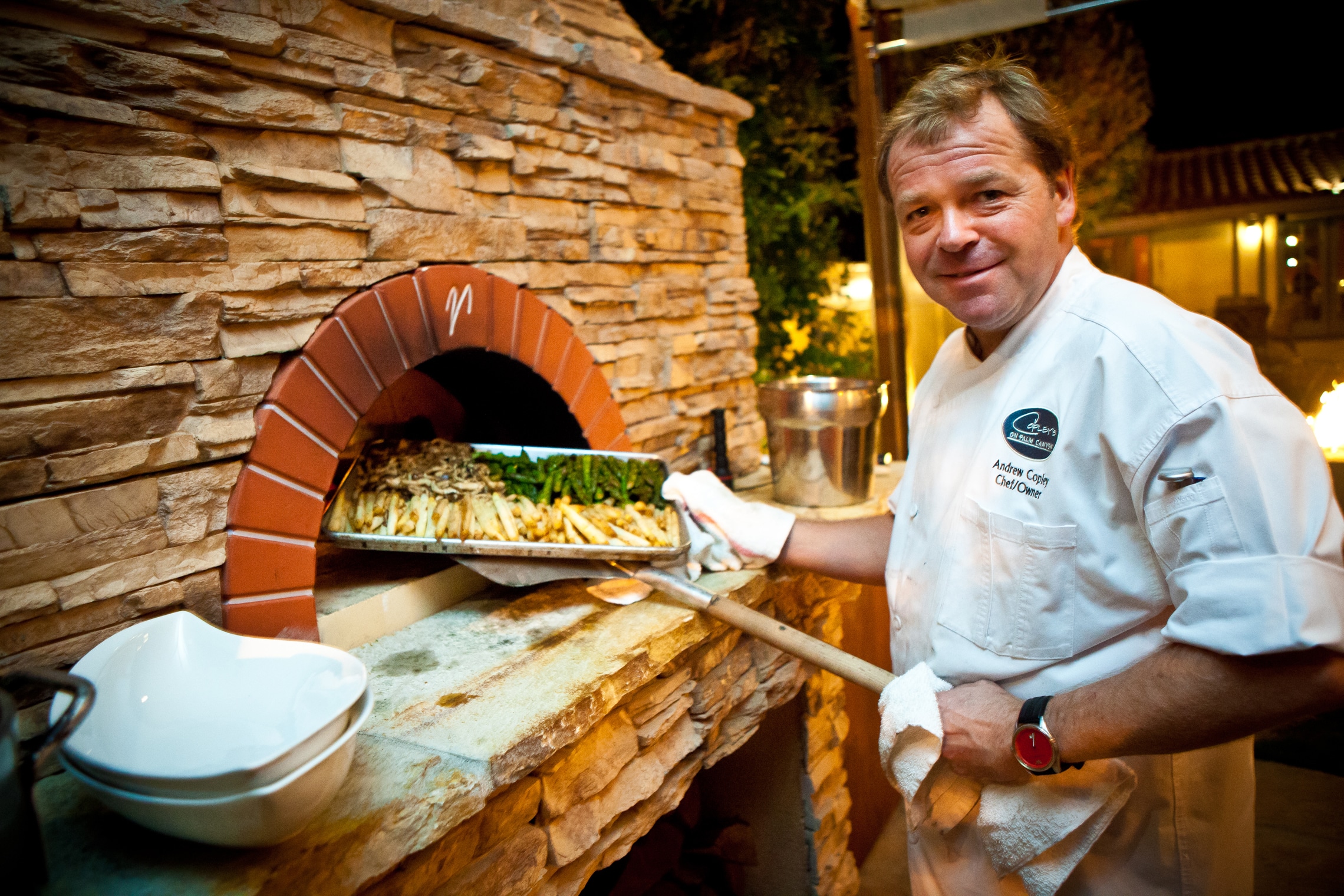 A chef smiles at the camera while pulling a pizza out of a stone pizza oven